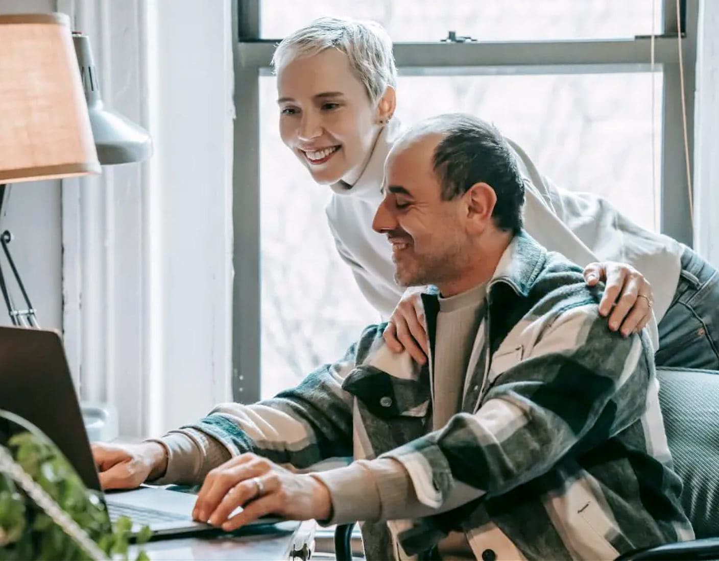 A smiling man sits at a desk with a woman standing behind him, both looking at a laptop while browsing for insurance options.